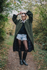 Person wearing a green coat, black sweater, denim shorts, and rain boots in a forest setting.