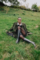 Person sitting on a grassy hill wearing a green coat, white shirt, and gray boots.