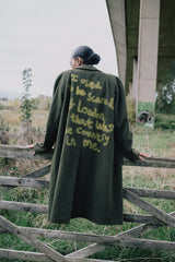 Person wearing a green coat with text, standing on a wooden fence in an outdoor setting.