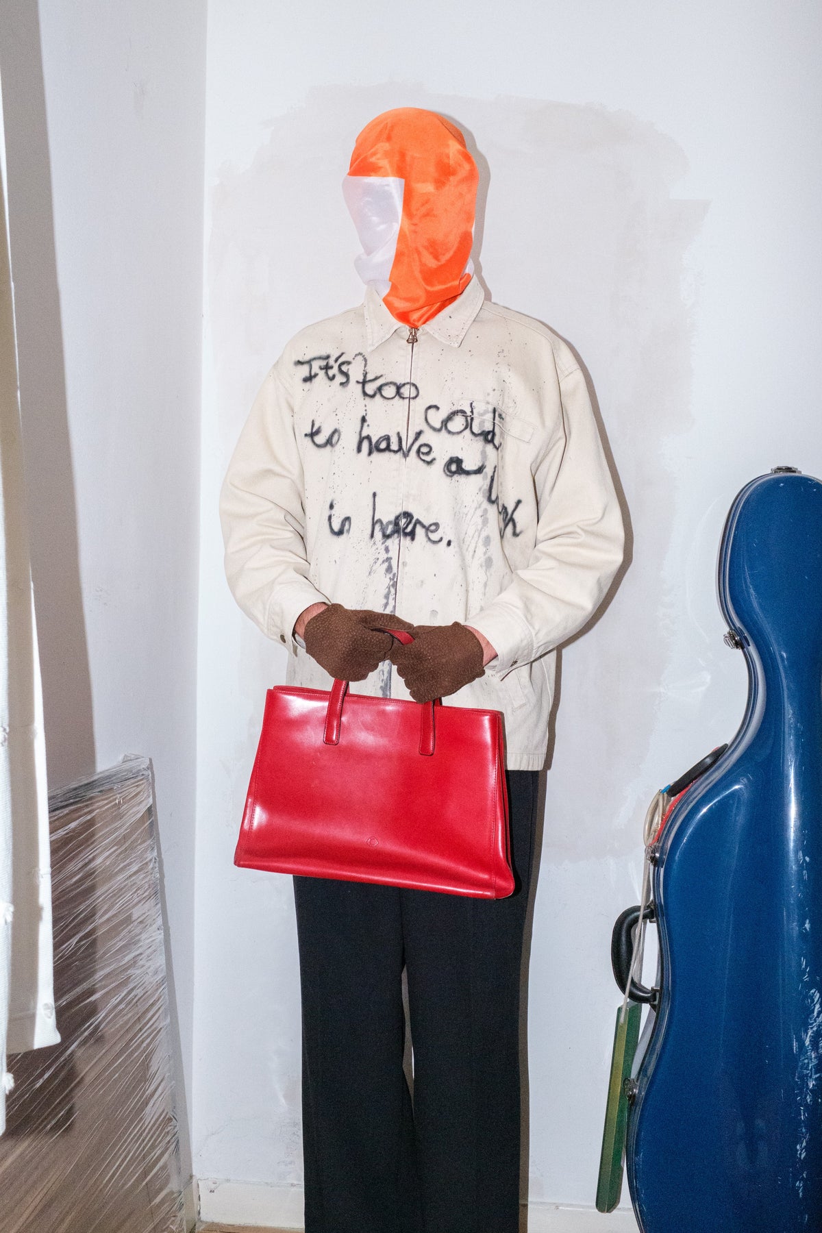 Mannequin wearing a beige jacket with text, holding a red bag, and standing next to a blue cello case against a white wall.