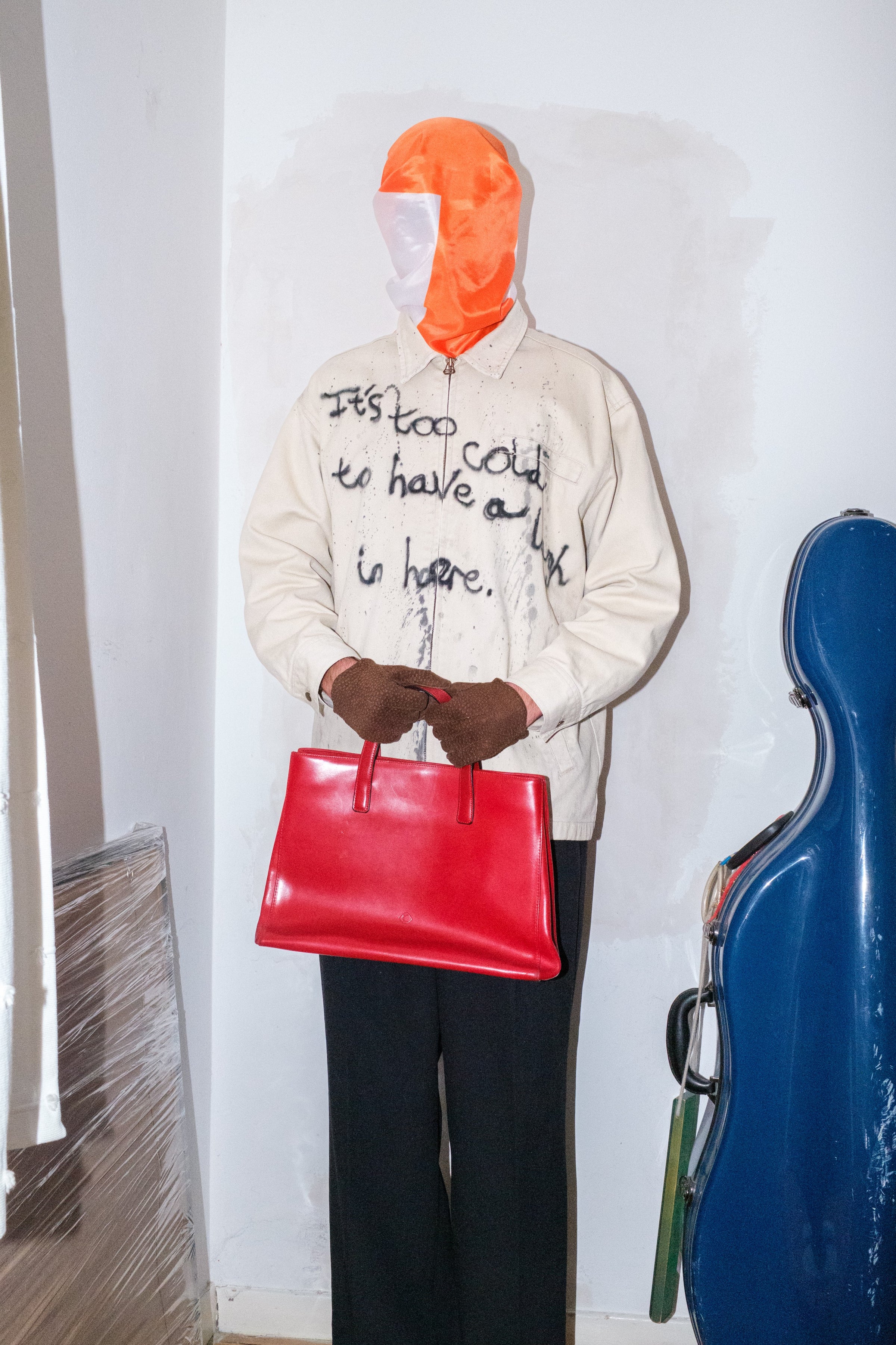 Mannequin wearing a beige jacket with text, holding a red bag, and standing next to a blue cello case against a white wall.