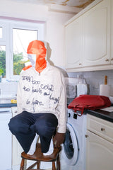Person sitting on a chair in a kitchen wearing a sweater with text and an orange mask.