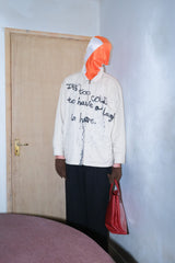 Person wearing a white jacket with text, orange hood, and dark pants standing in a room with a door and light blue wall.