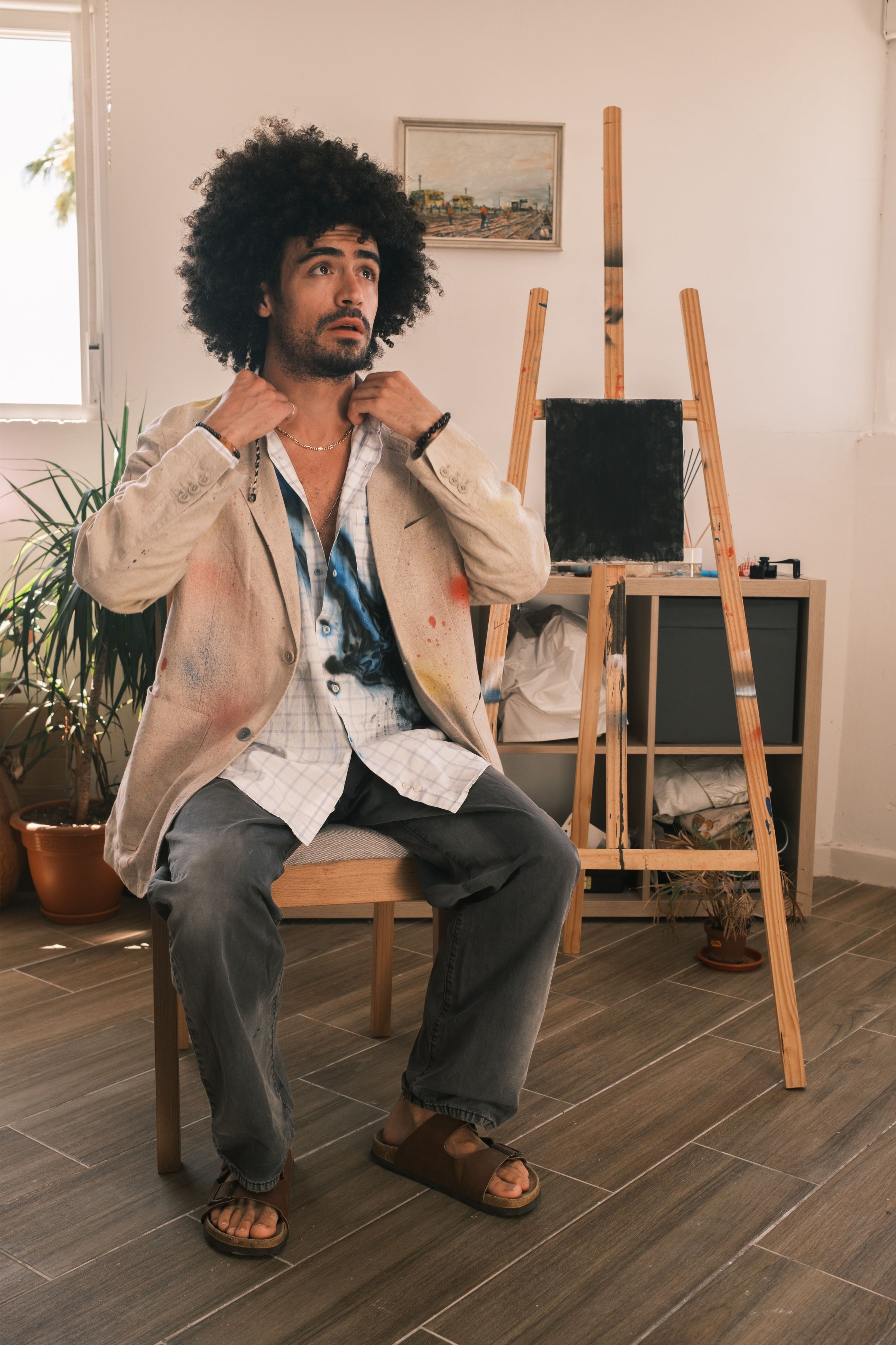Model seated indoors wearing beige blazer over spray-painted check shirt, art studio setting, Bewept Bag of Man.